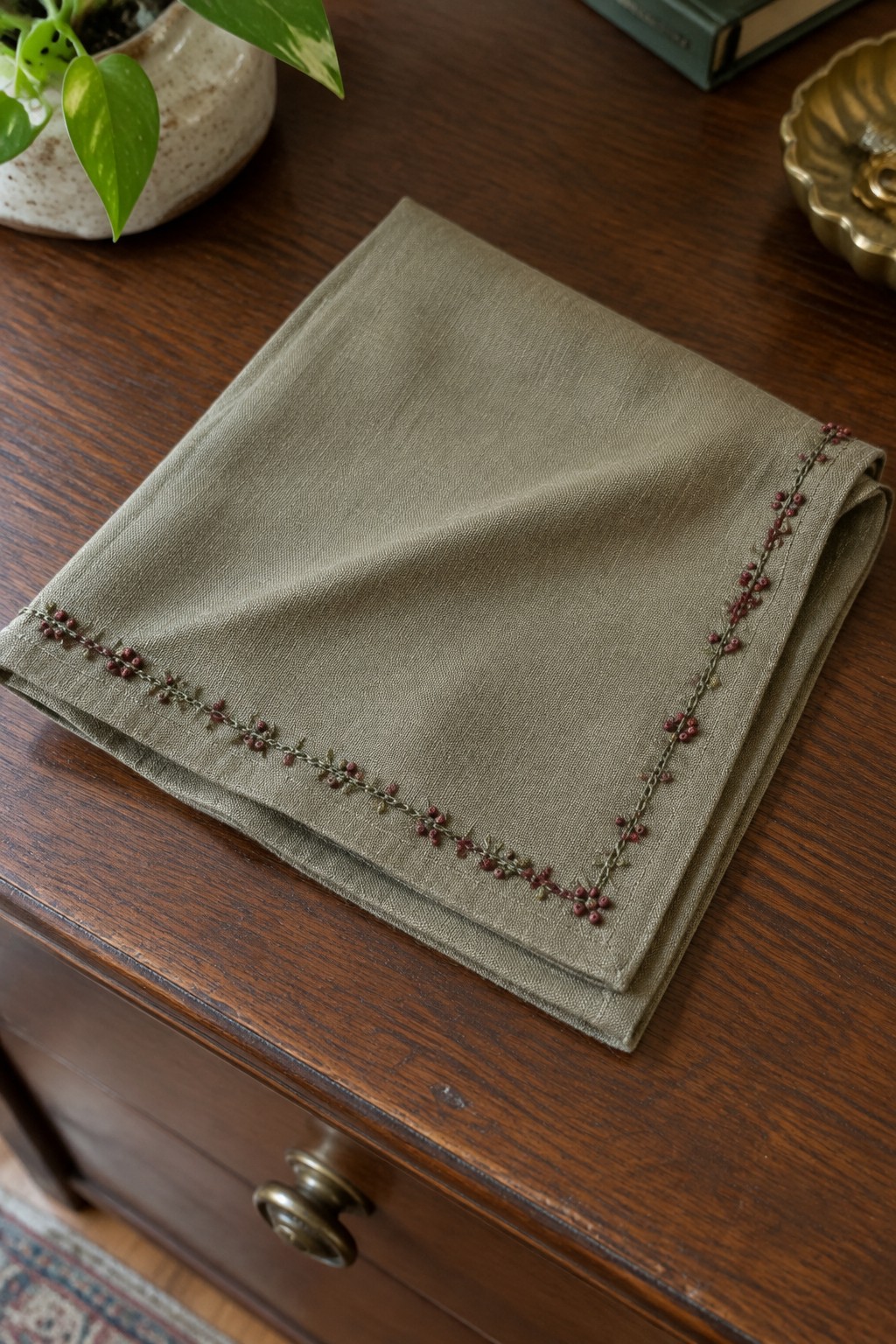 A folded taupe linen napkin with a stitched green vine and dark red berry border rests on a wooden table.