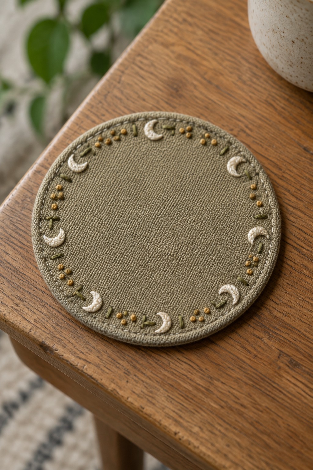 A round olive fabric mat embroidered with cream crescent moons, gold dots, and small leaf-like marks around the edge.