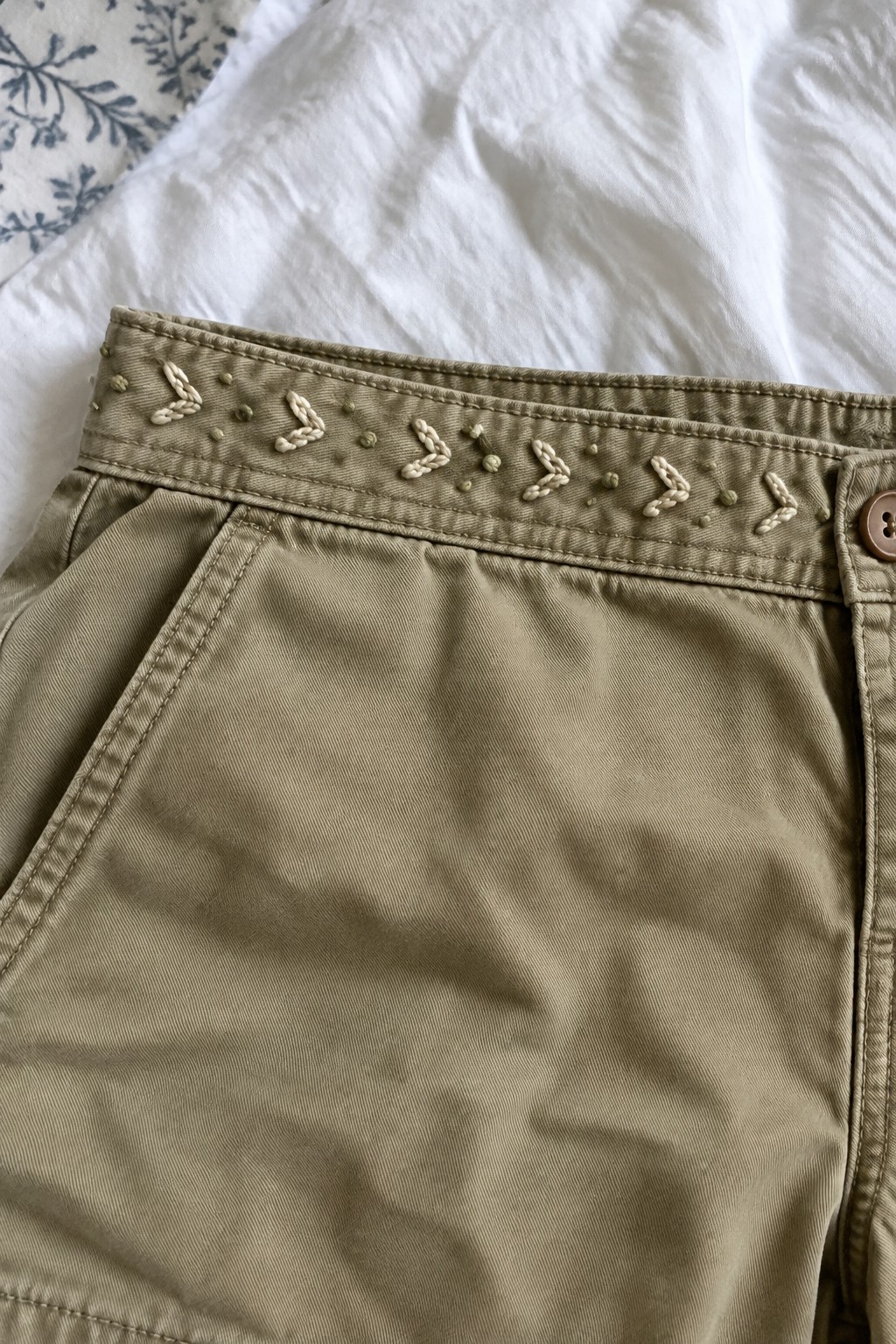 Khaki trousers with a cream repeating chain motif embroidered along the waistband.