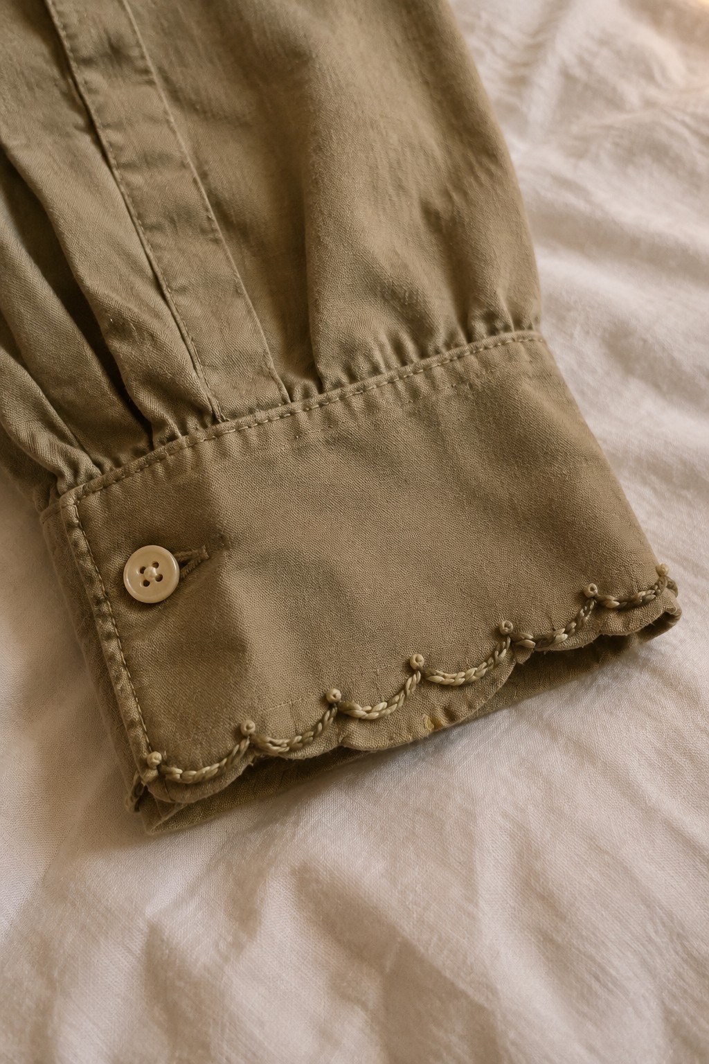 A khaki-colored shirt cuff with a button and a looped scallop border lies on white fabric.