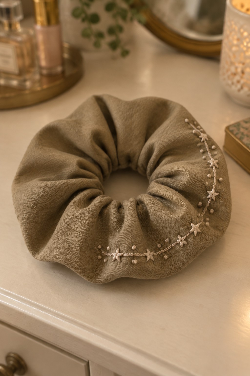 A taupe fabric scrunchie with a curved embroidered border of small flowers and dots rests on a cream vanity.