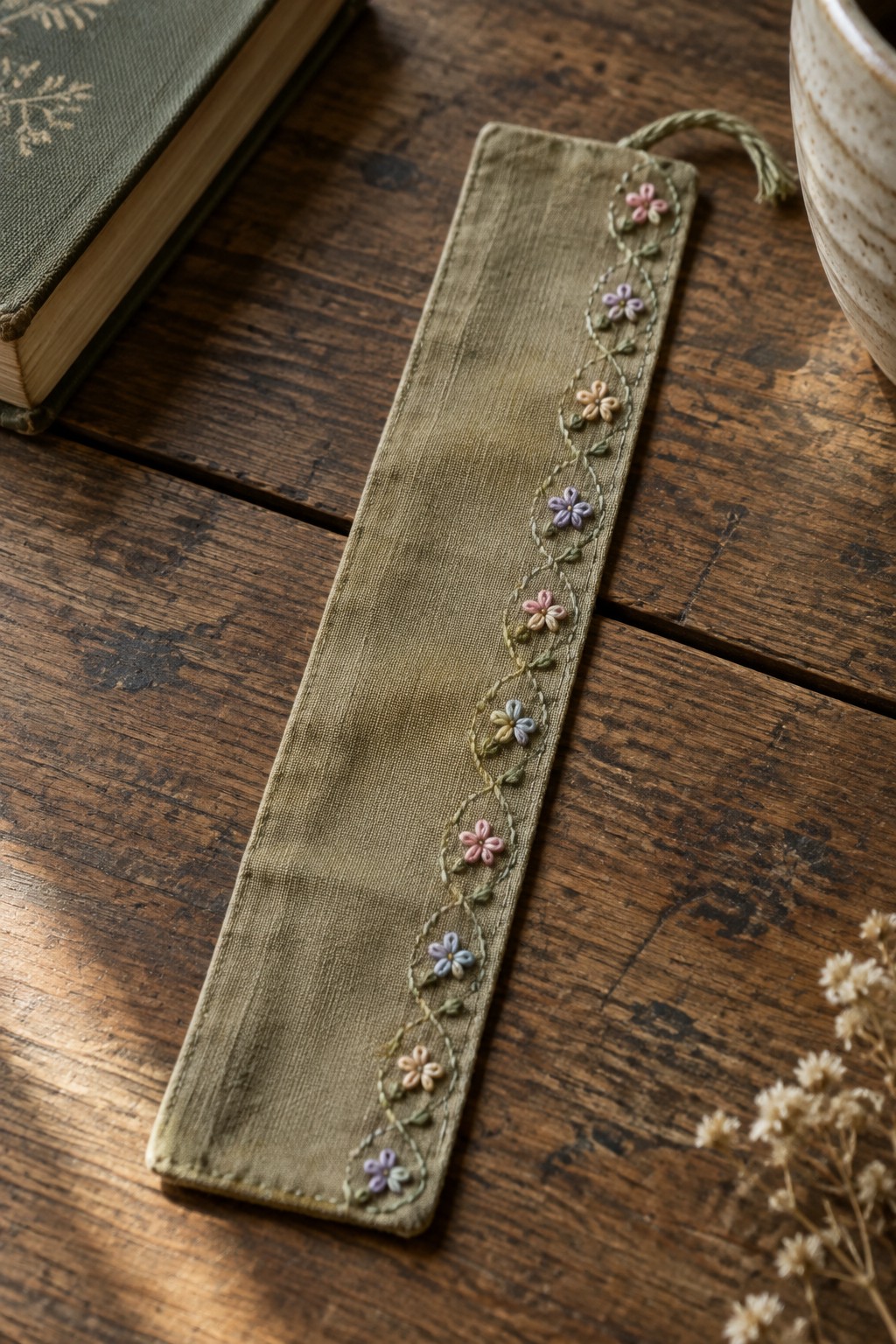 A khaki fabric bookmark with a stitched border of small pastel flowers and leaves on a wooden table.