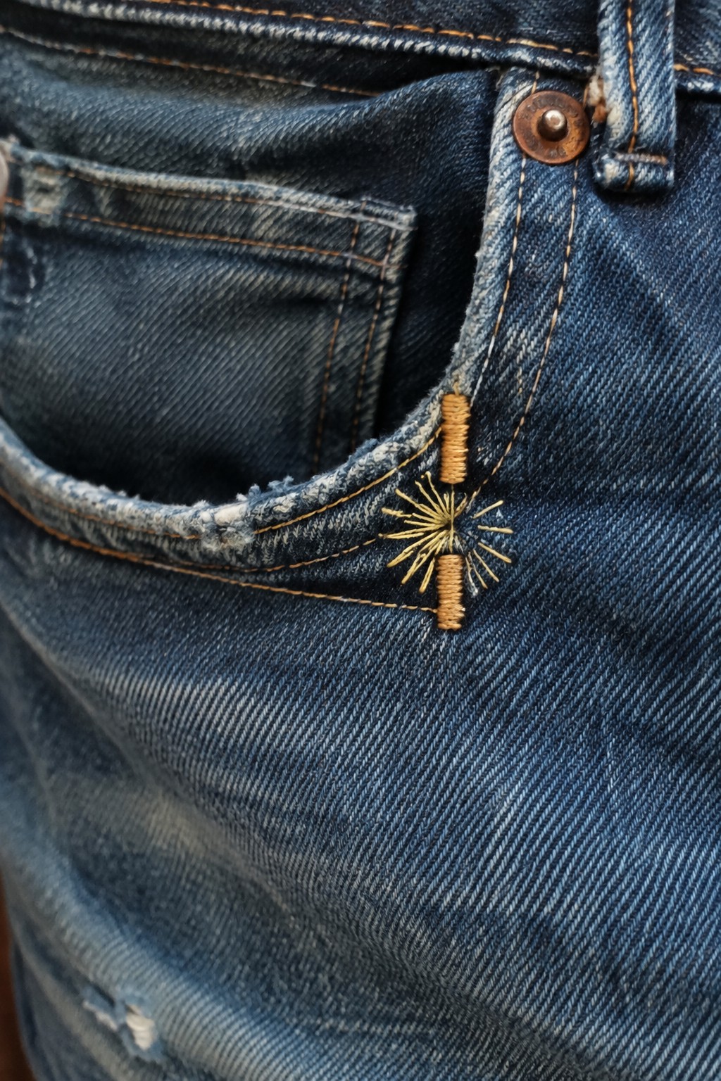 Close-up of blue jeans with a golden sunburst embroidered beside a frayed pocket edge.