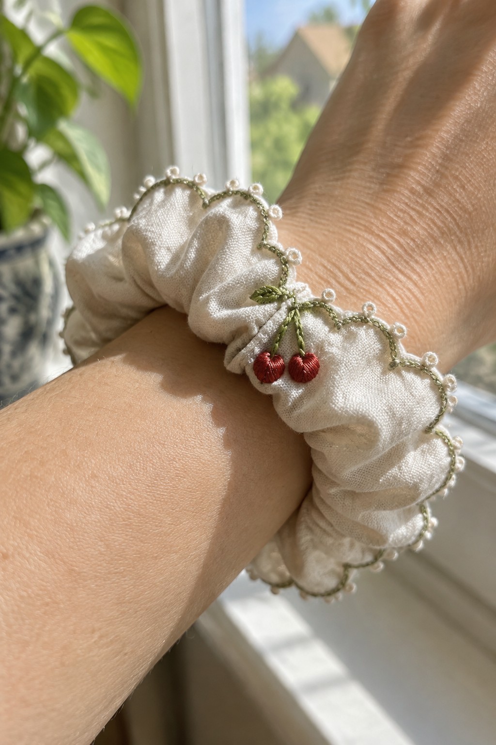 A white gathered scrunchie with a green scalloped embroidered border and two red cherries worn on a wrist.