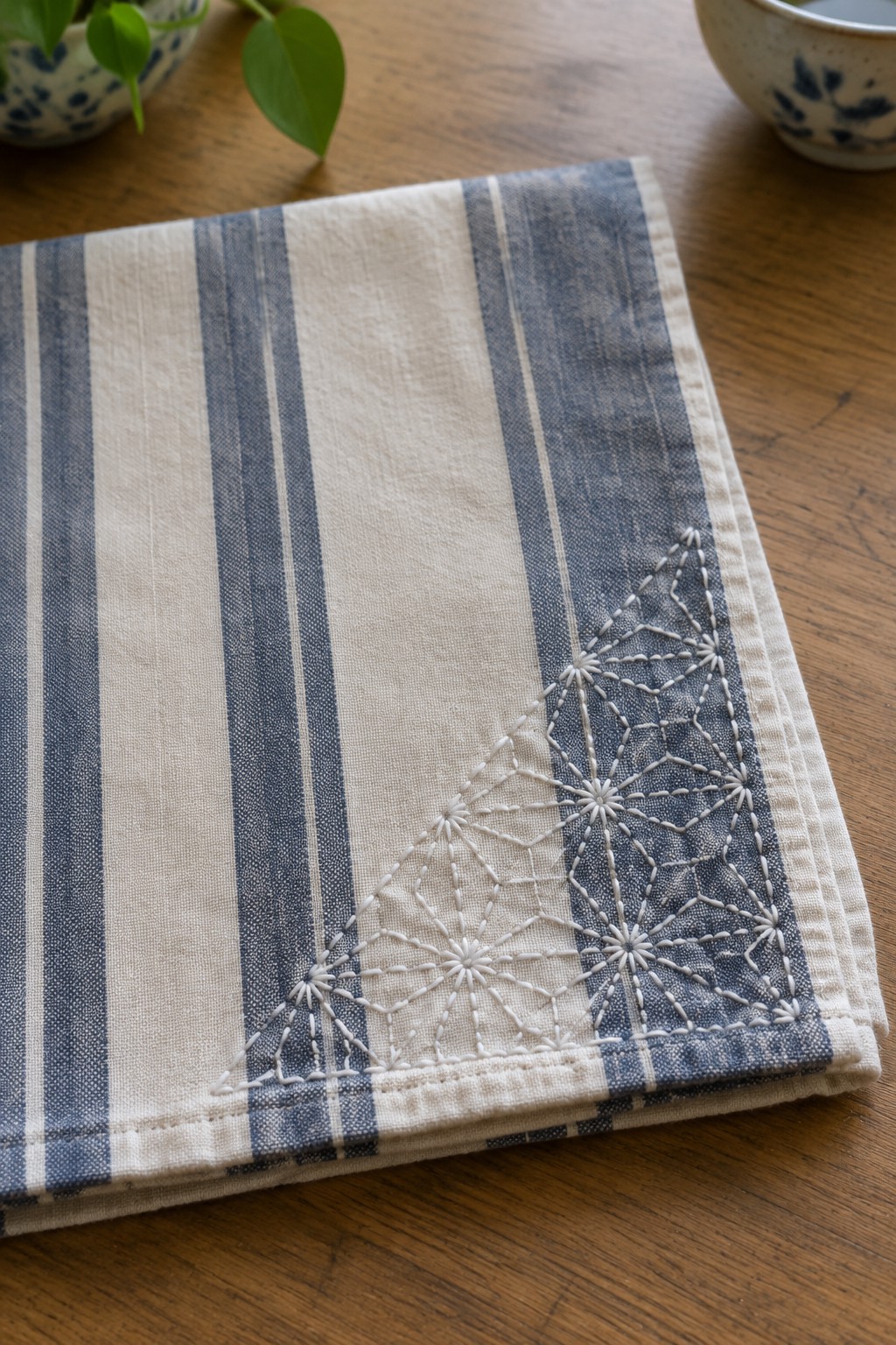 White geometric hemp-leaf embroidery decorates the corner of a blue-and-cream striped tea towel on a wooden table.
