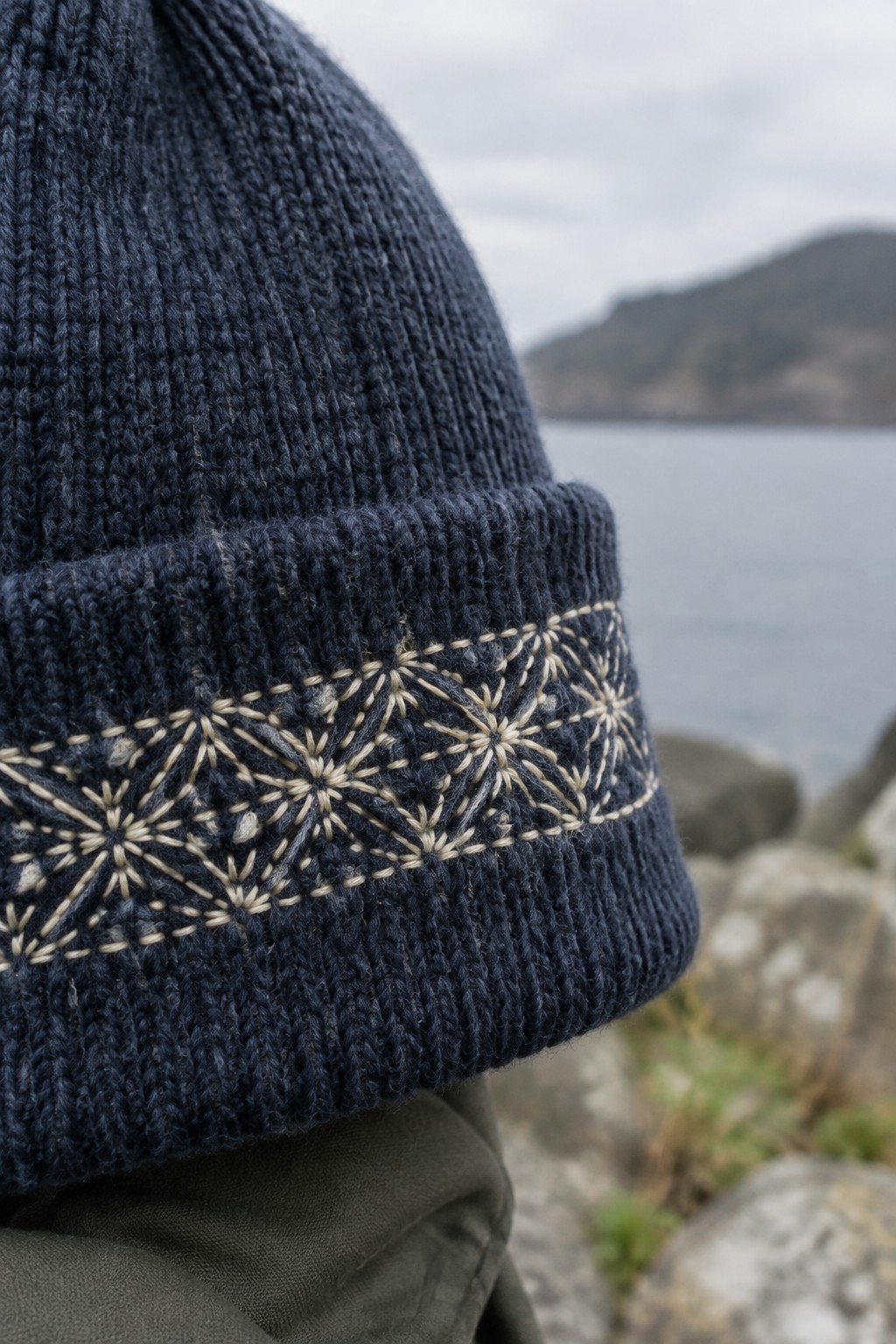 A dark navy knit beanie with a cream asanoha embroidery band on its folded cuff, photographed beside a rocky shoreline.