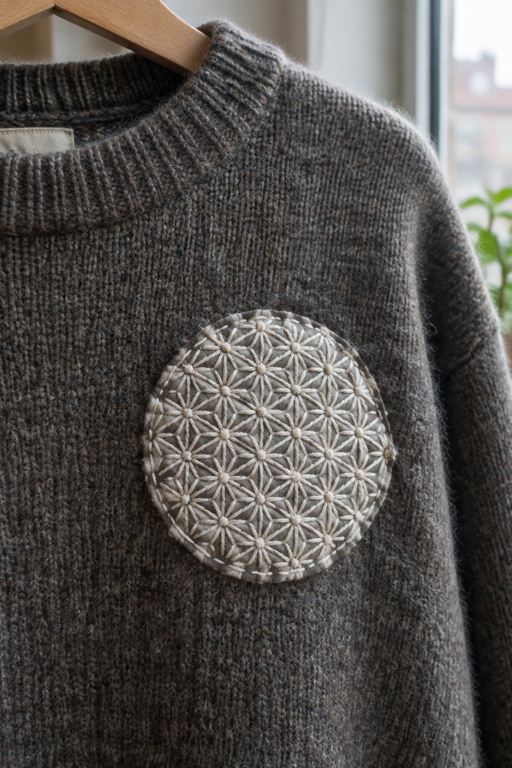 A circular cream asanoha embroidery patch is pinned to the chest of a gray-brown knitted sweater on a wooden hanger.