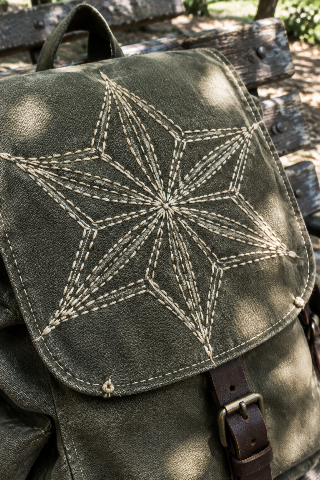 Cream geometric stitching decorates the flap of an olive fabric backpack resting against a wooden bench.