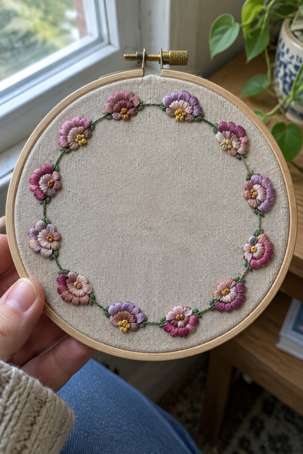 A wooden embroidery hoop holds a circular border of pink and purple flowers stitched on beige fabric.