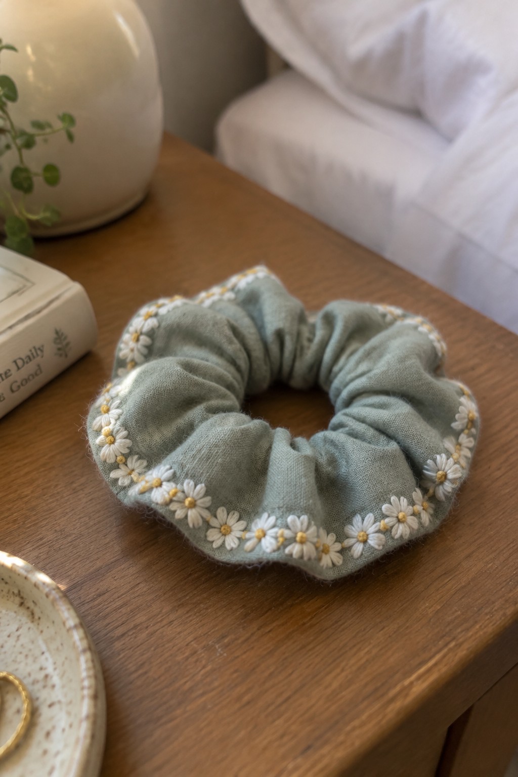 A pale green scrunchie with white and yellow embroidered daisies rests on a wooden table.