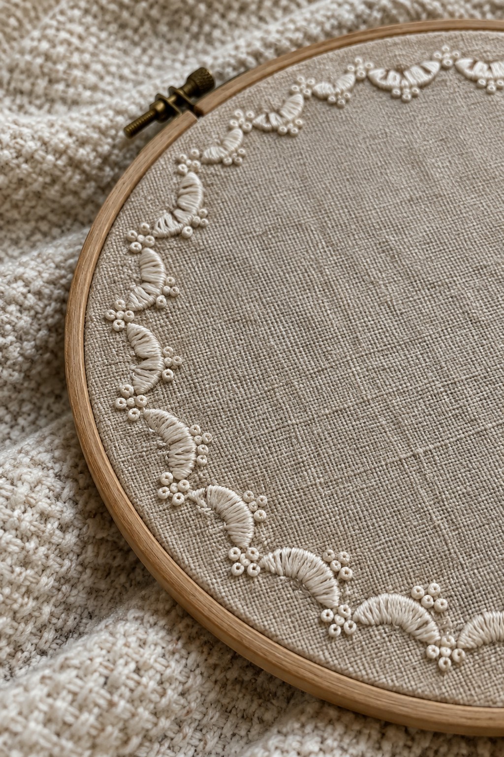 Close-up of a cream embroidered border with raised thread motifs and small bead-like clusters around the edge of linen stretched in a wooden hoop.