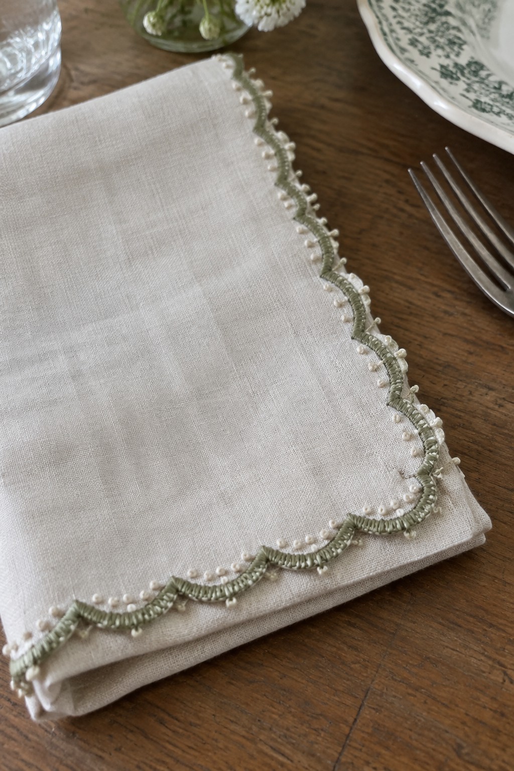 A folded cream linen napkin with a sage green scalloped embroidered border and small white accents rests on a wooden table.