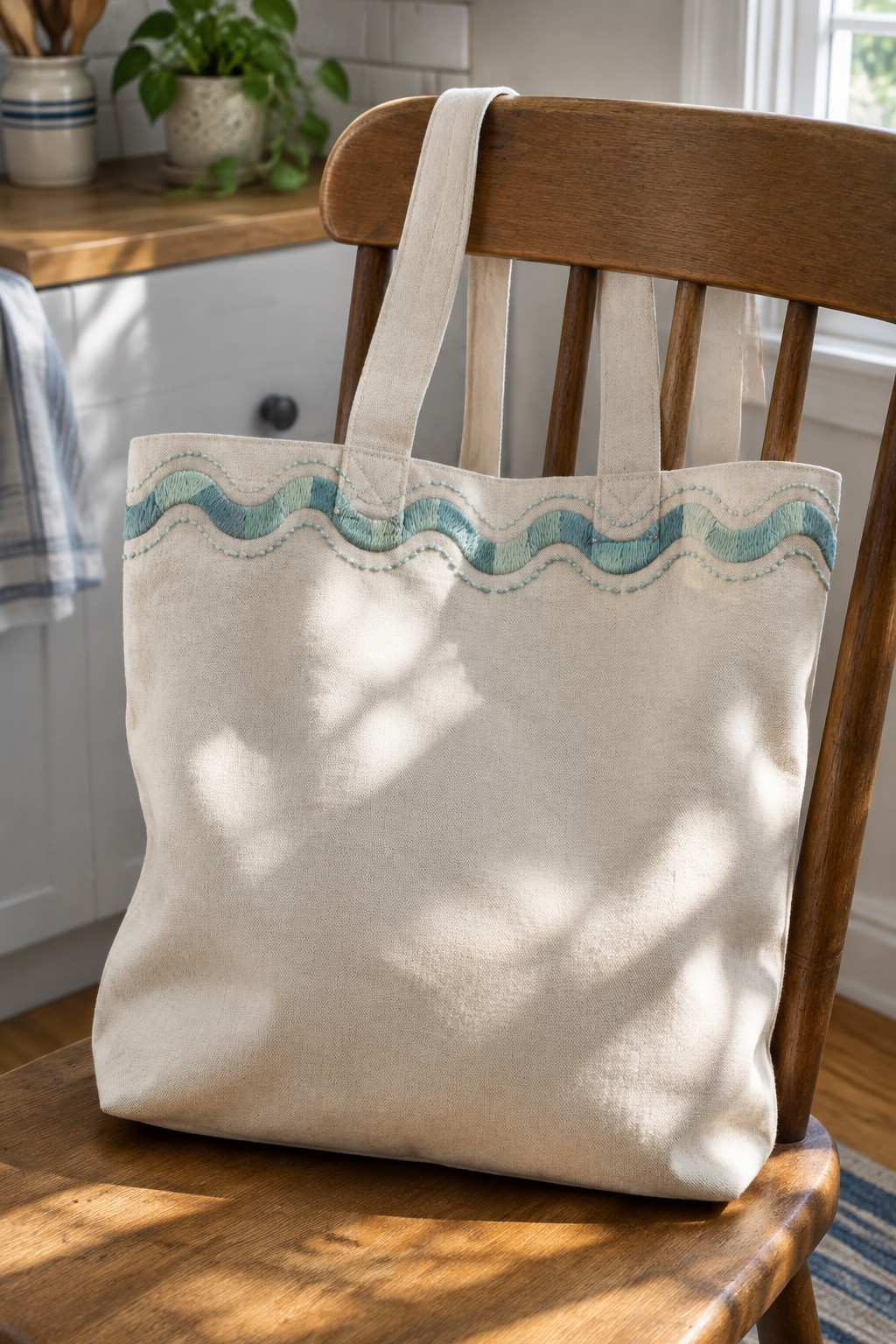 An ivory canvas tote with a teal wavy embroidered border hanging on a wooden chair.