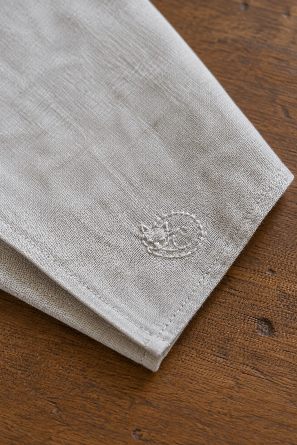A small white cat outline embroidered near the hemmed corner of a folded beige linen napkin on a wooden table.