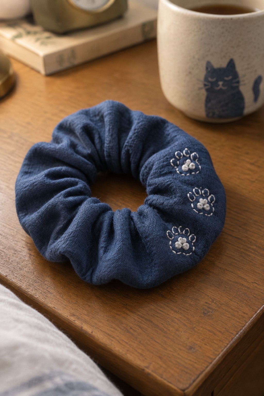 A navy gathered scrunchie embroidered with three white paw prints rests on a wooden table beside a cat-print mug.