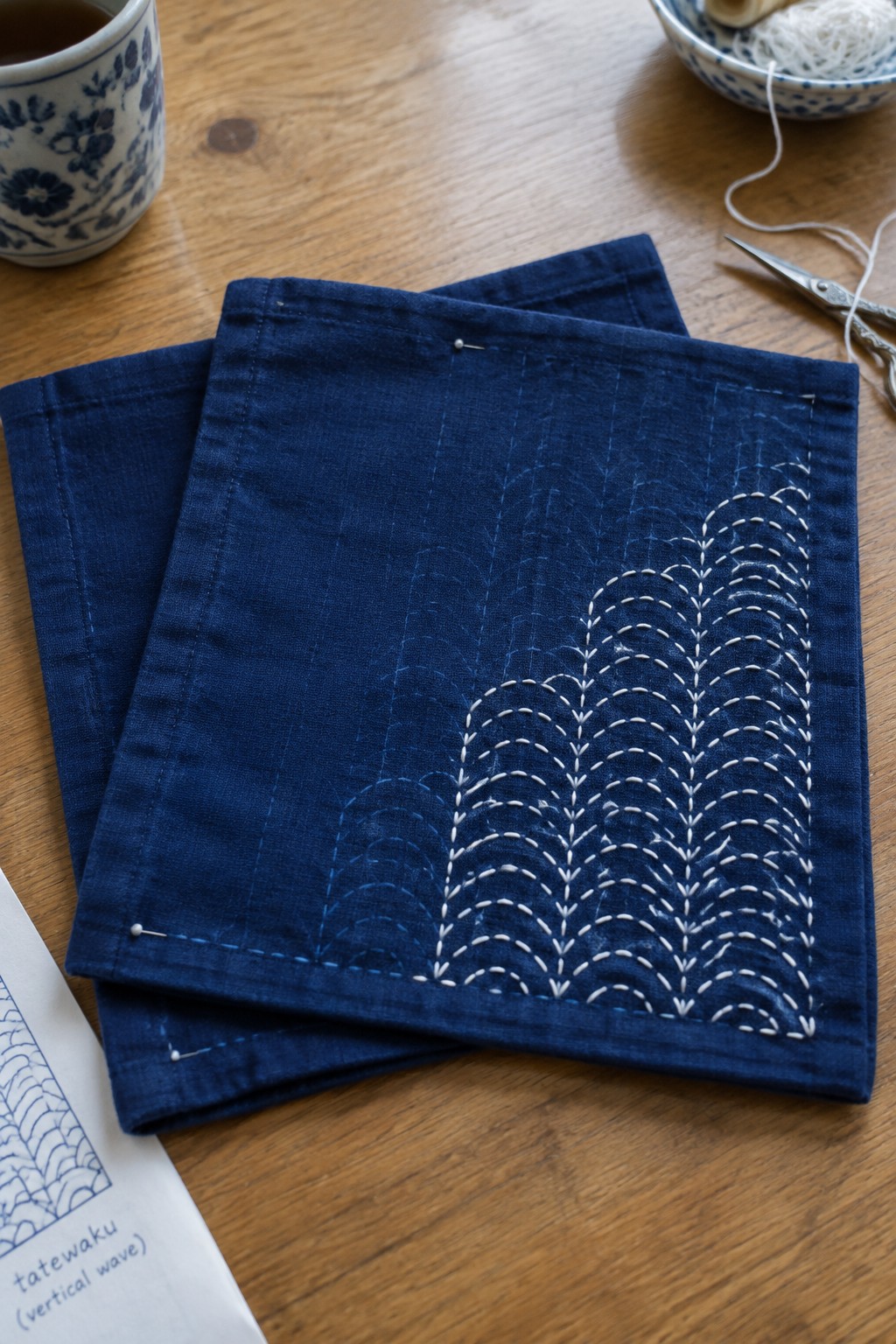 White stitched wave patterns cover one corner of a dark blue fabric panel pinned over two matching panels on a wooden table.