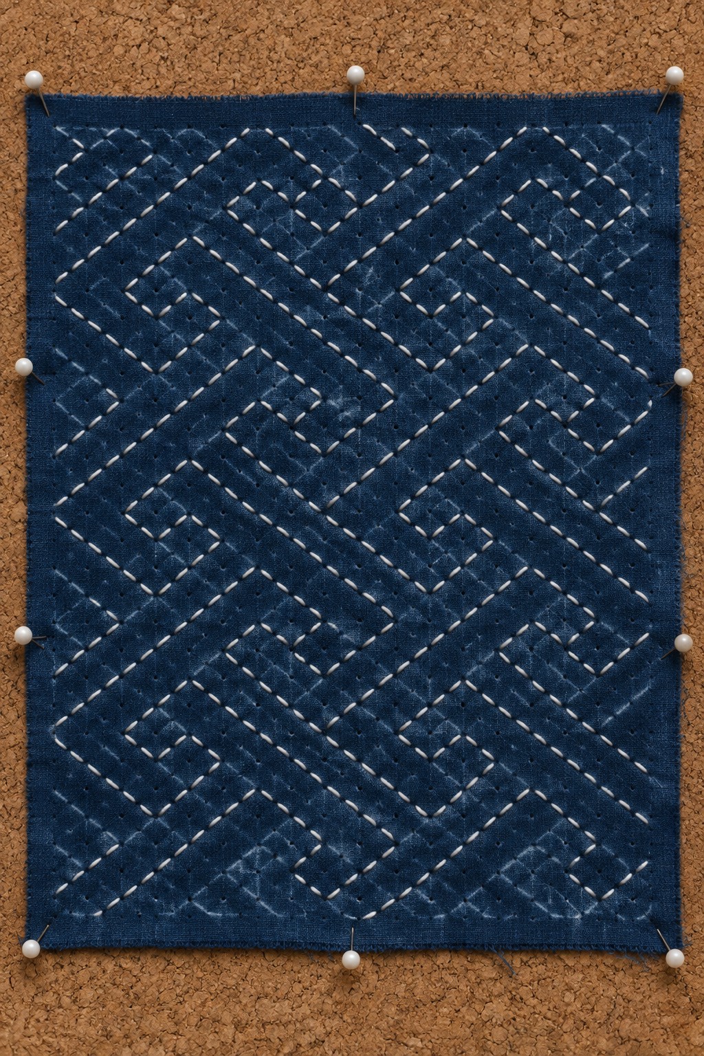 White geometric running stitches form a maze pattern across a rectangular indigo fabric panel pinned to cork.