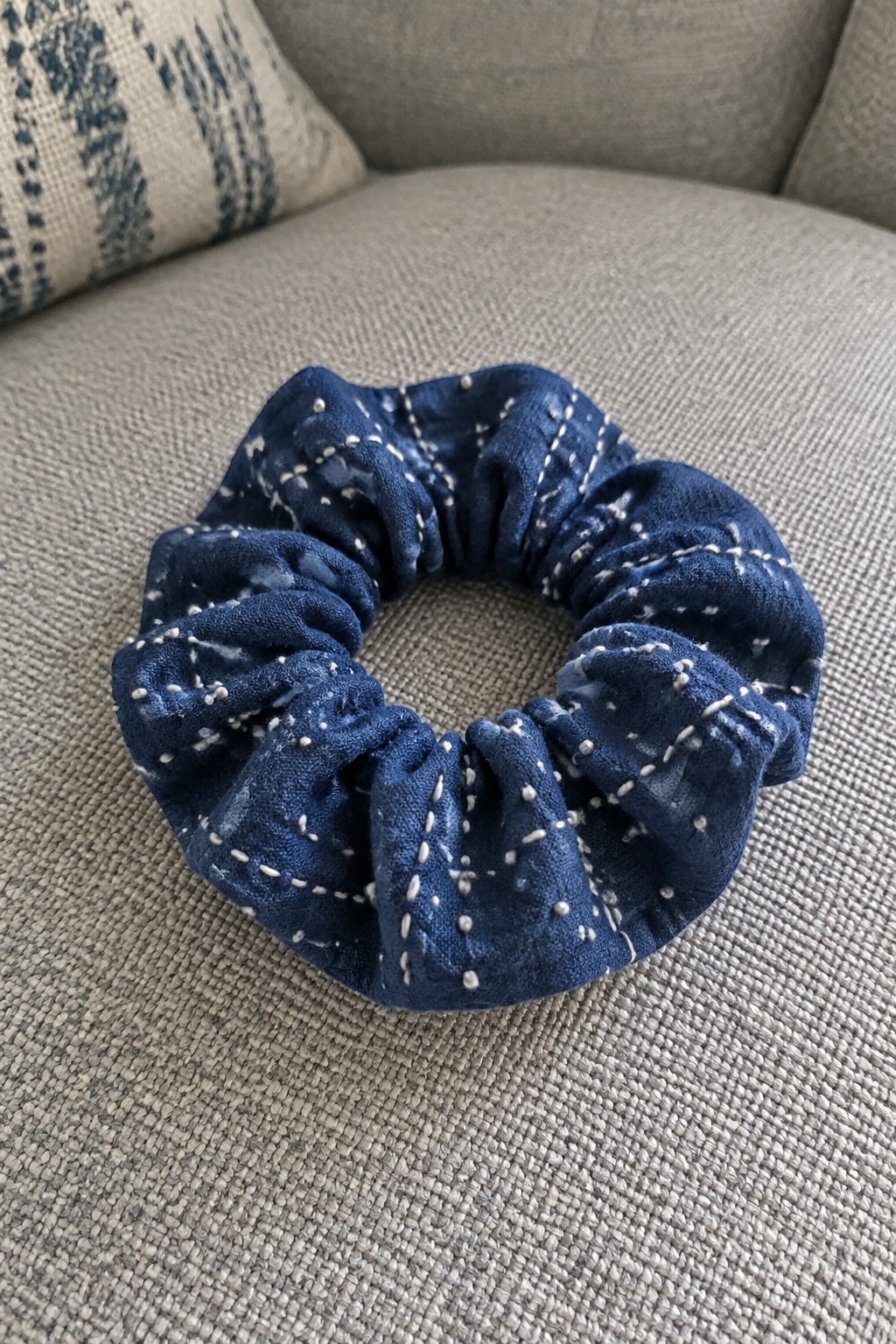 A navy fabric scrunchie with pale stitched lines and small marks rests on a gray sofa.