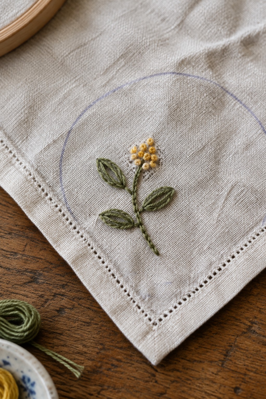 A small yellow embroidered flower with green leaves sits on a linen handkerchief inside a penciled circle.