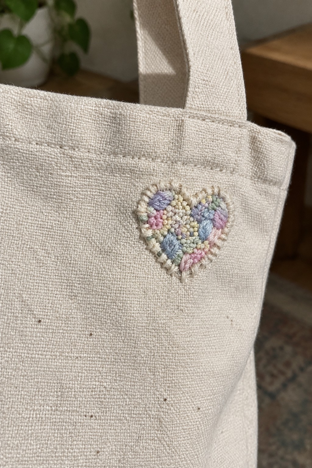 A cream canvas tote with a small textured pastel heart embroidered on the front near the top edge.