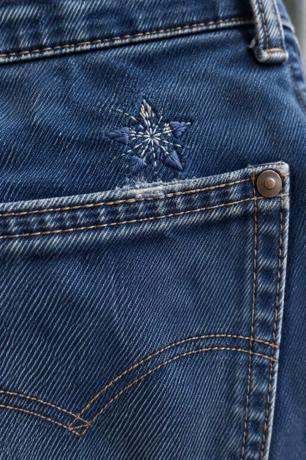 A small blue and white embroidered flower sits above the pocket seam on dark blue jeans.