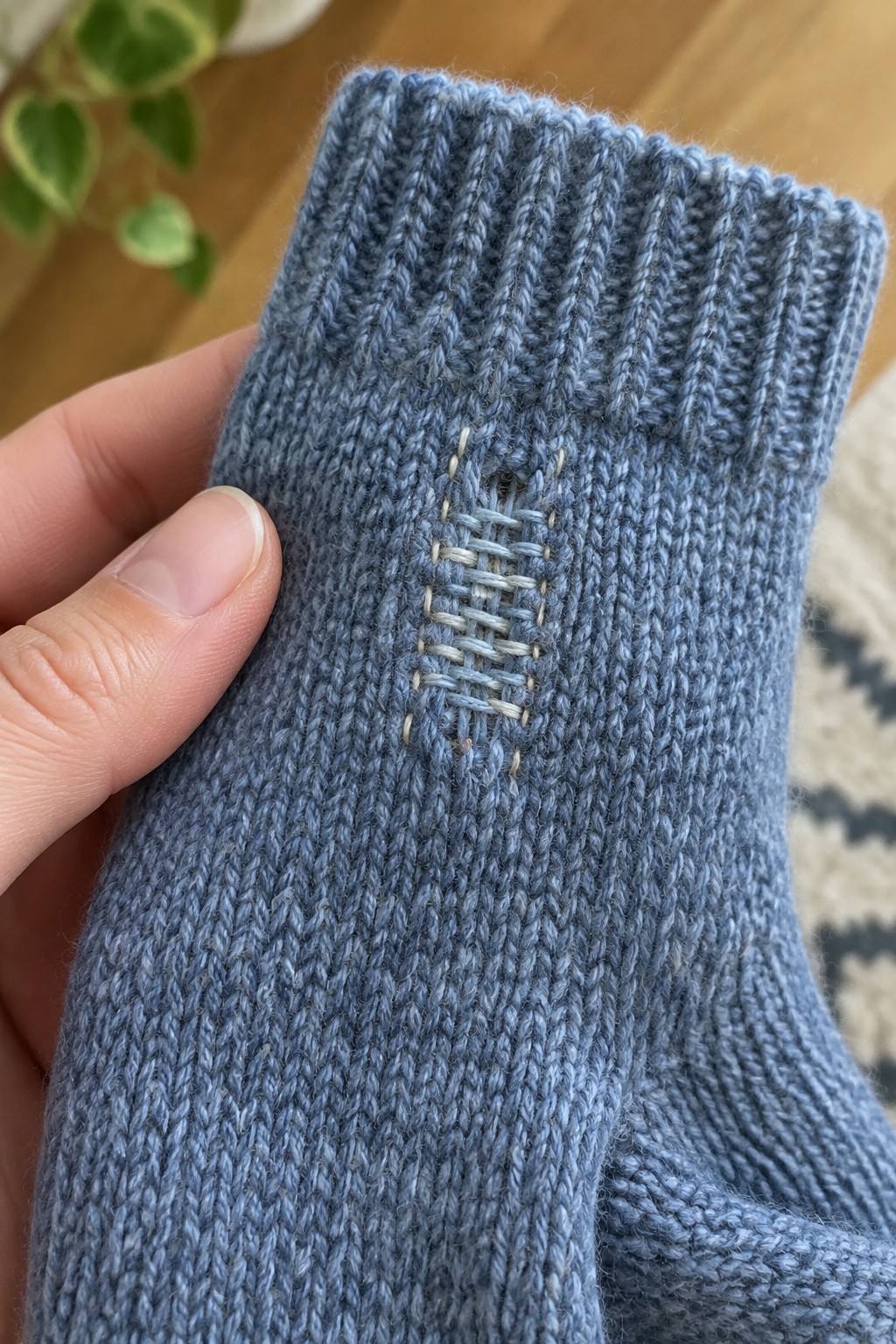A blue knitted sock held in a hand, with a small hole mended using pale woven stitches.