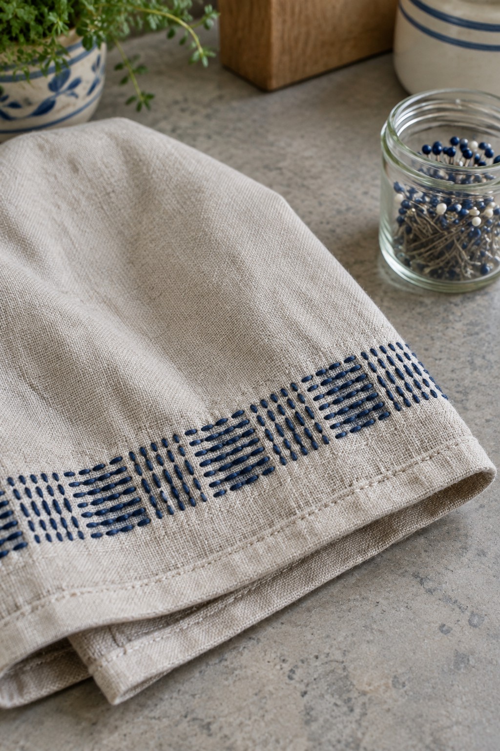 A beige linen towel with a blue geometric stitched border lies on a stone countertop beside a jar of pins.