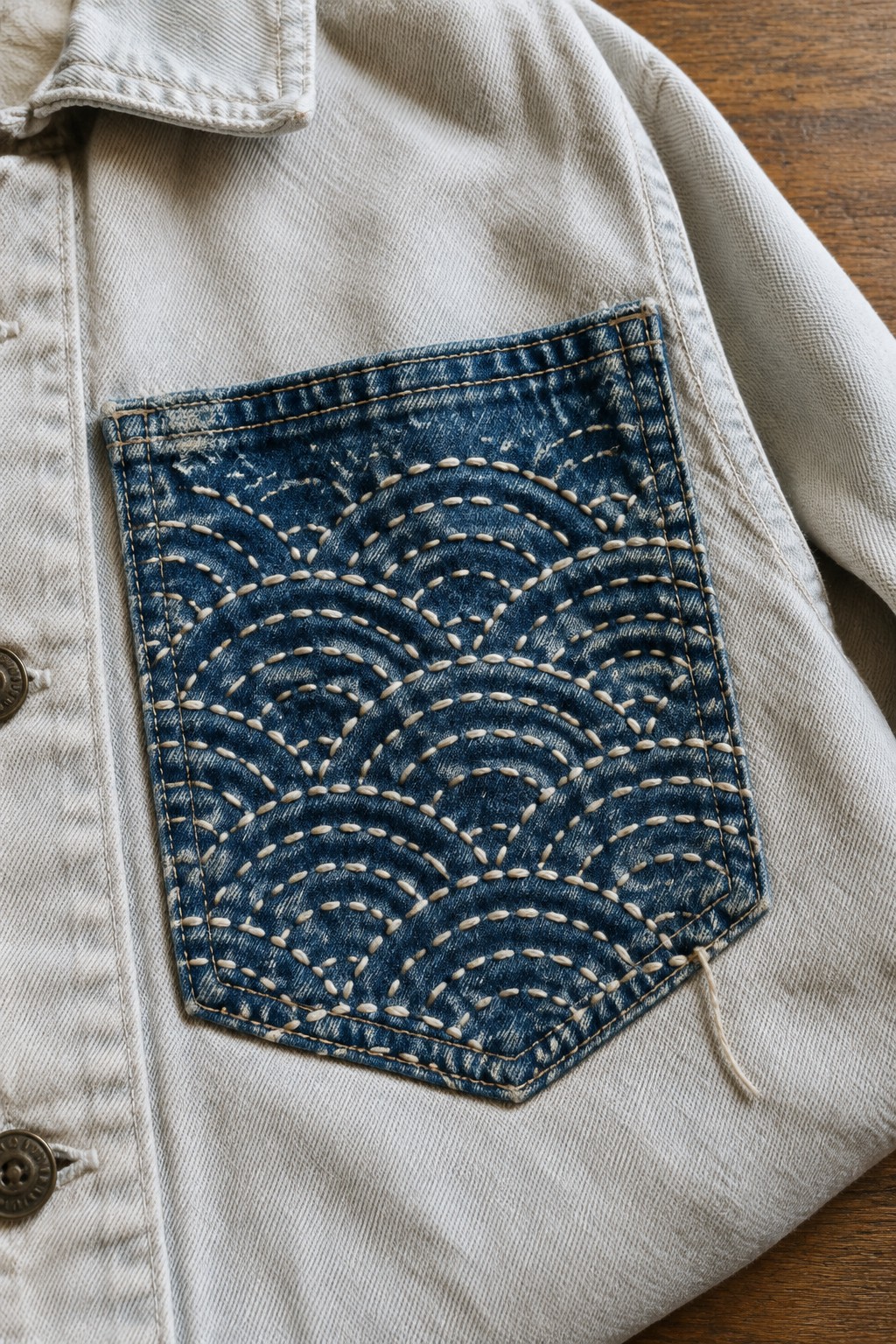 A dark denim pocket with pale curved sashiko-style stitching sewn onto a light jacket.