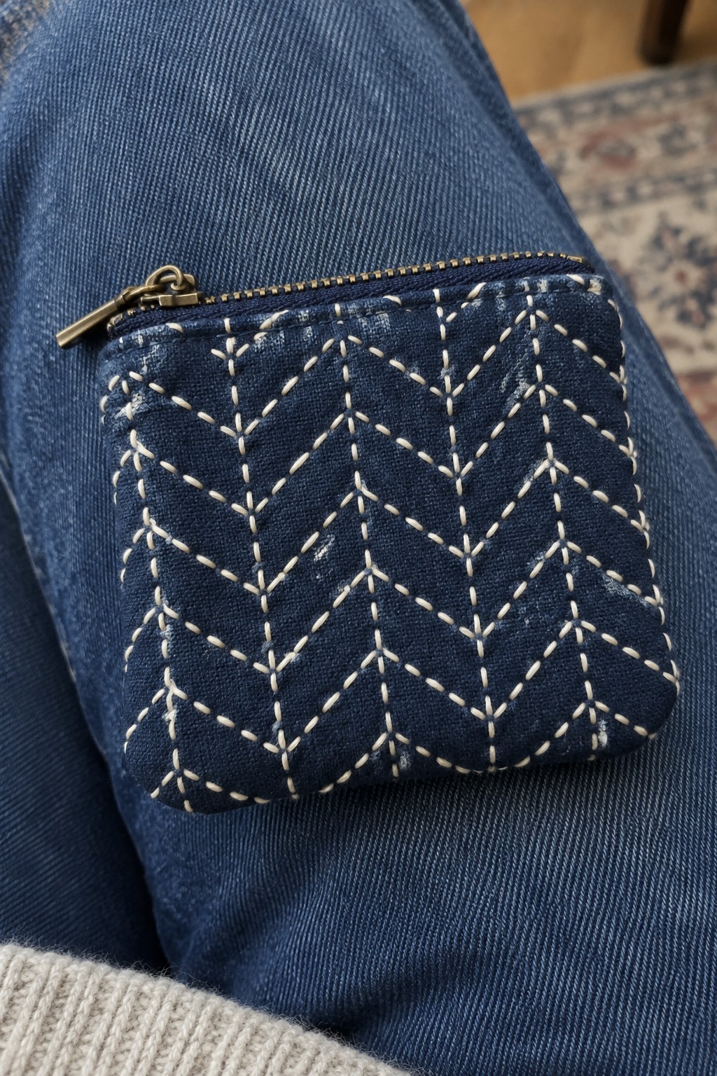 A navy zippered pouch embroidered with a cream geometric lattice rests on blue jeans.