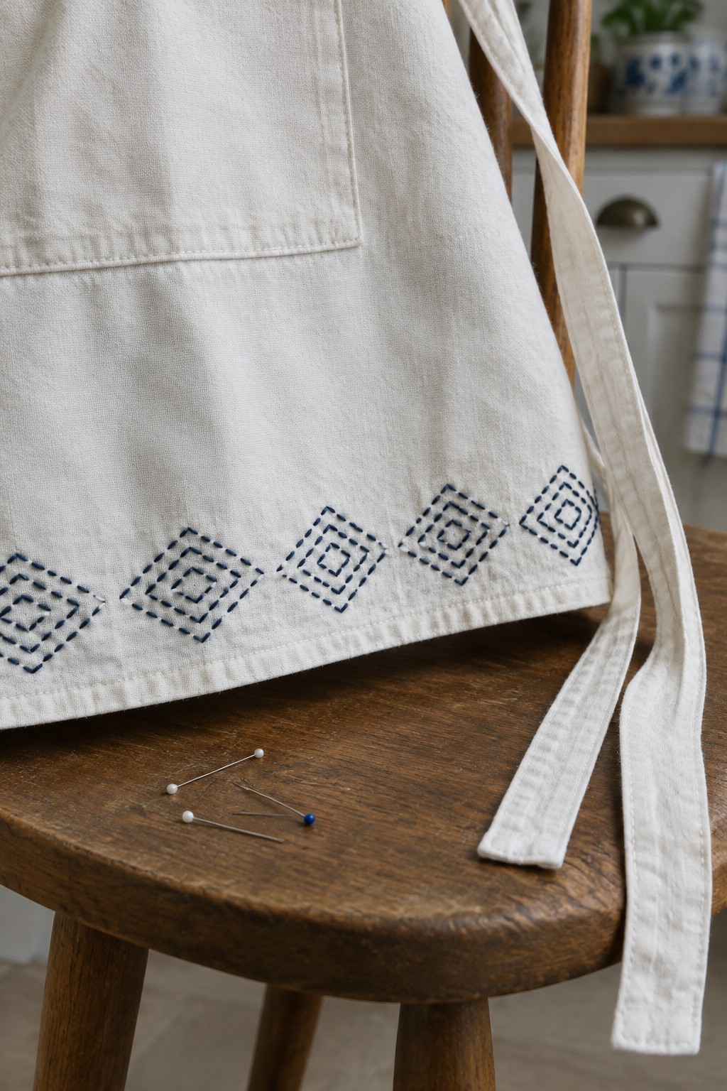 A cream apron with dark blue stitched diamond motifs along the lower hem rests on a wooden chair beside pins.