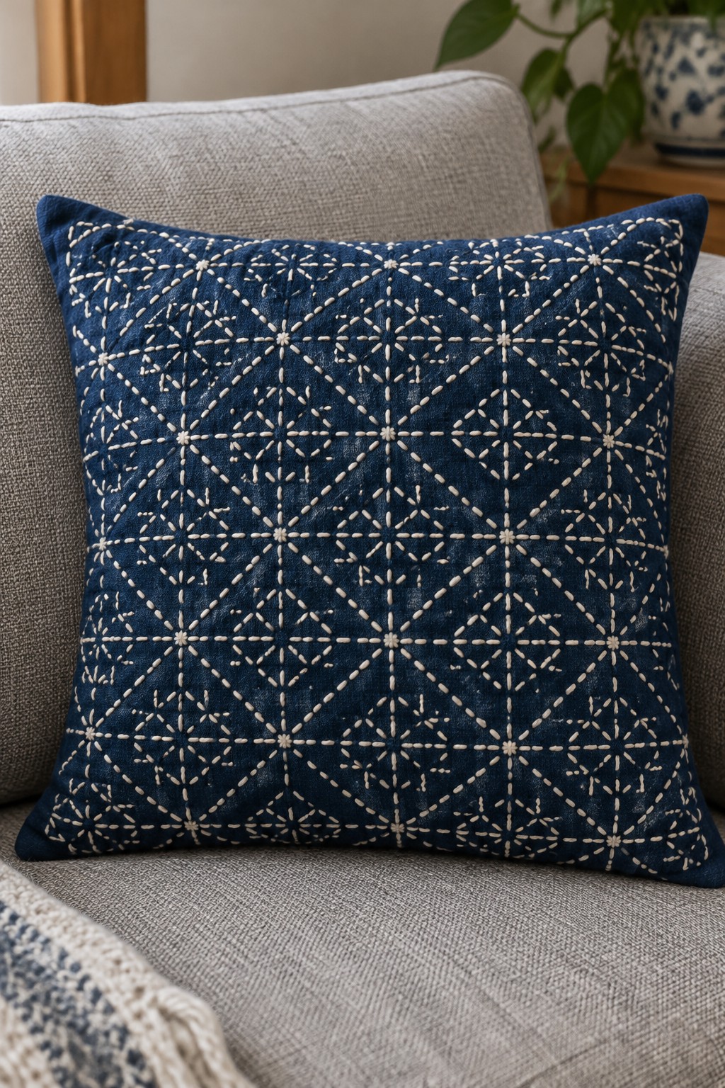 A navy cushion cover embroidered with a repeating cream geometric sashiko pattern rests on a gray sofa.