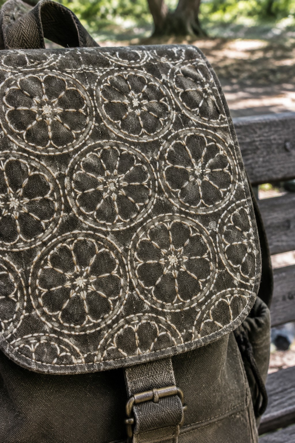 A dark olive backpack with cream circular floral embroidery across its flap rests against a wooden bench.
