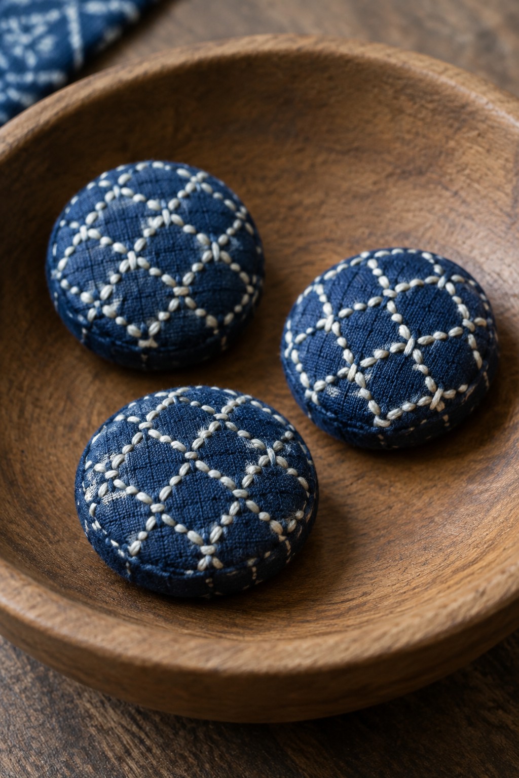 Three round indigo fabric buttons with cream geometric stitching sit in a wooden bowl.