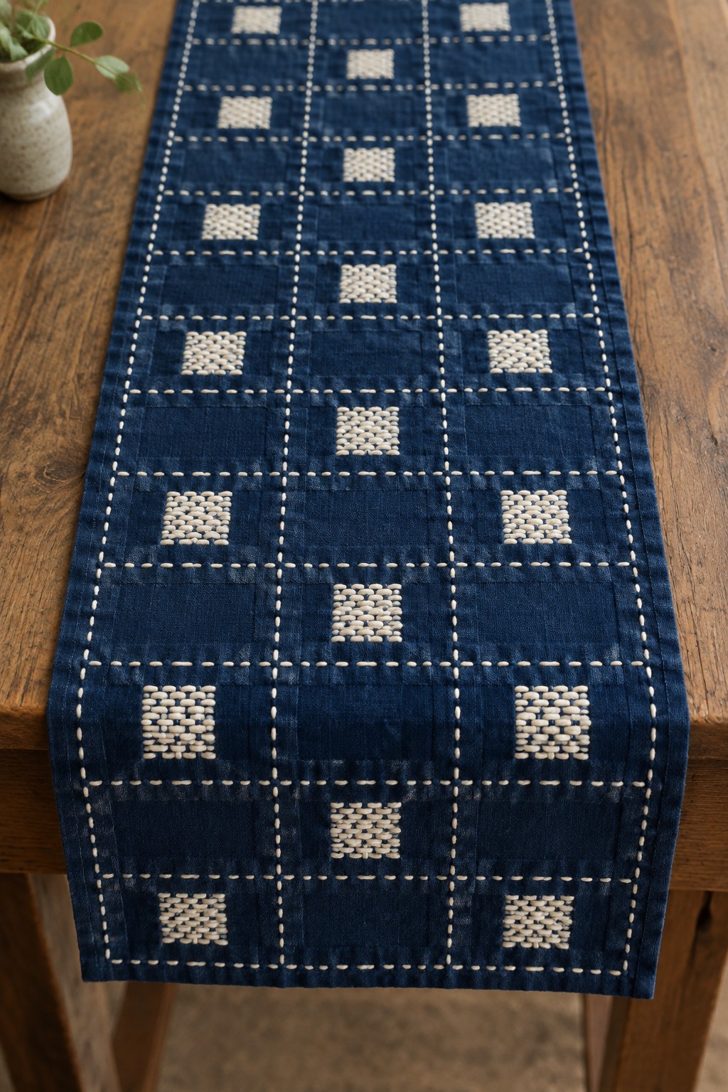 A deep blue table runner with a cream stitched grid and small cream textured squares lies across a wooden table.