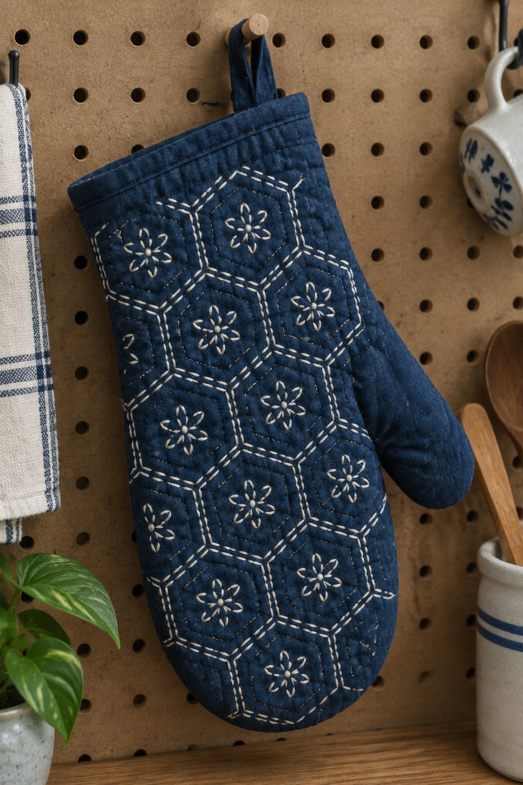 A dark blue oven mitt with cream sashiko-style hexagon and floral stitching hangs on a pegboard.