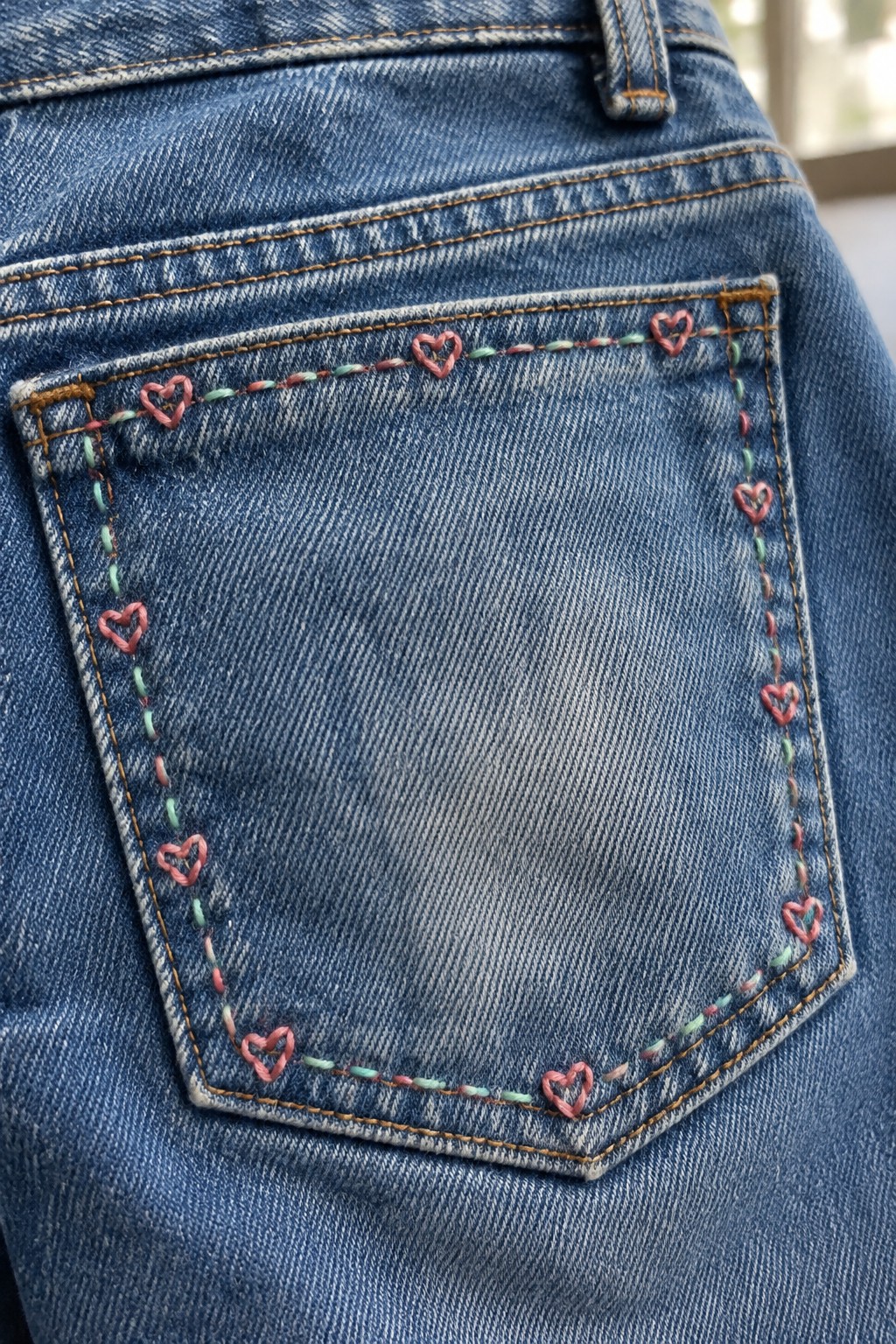 Pink heart outlines and alternating pink and green stitches decorate the edge of a blue denim back pocket.
