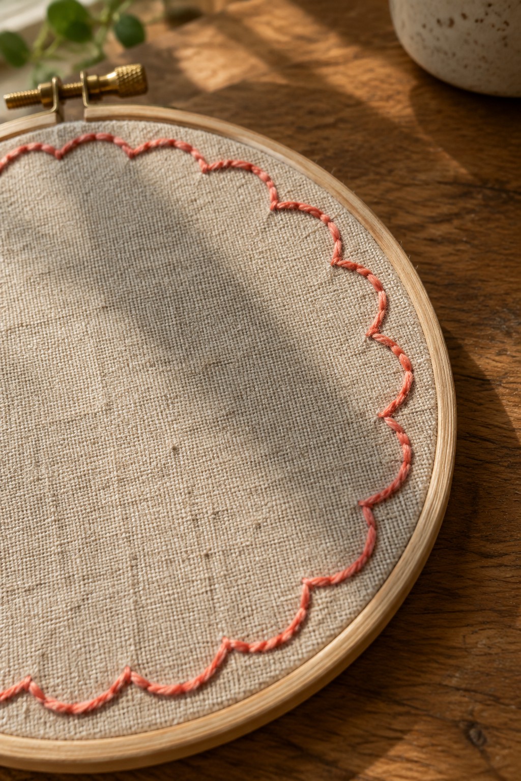 Coral scalloped stitching outlines the edge of beige fabric stretched in a wooden embroidery hoop.