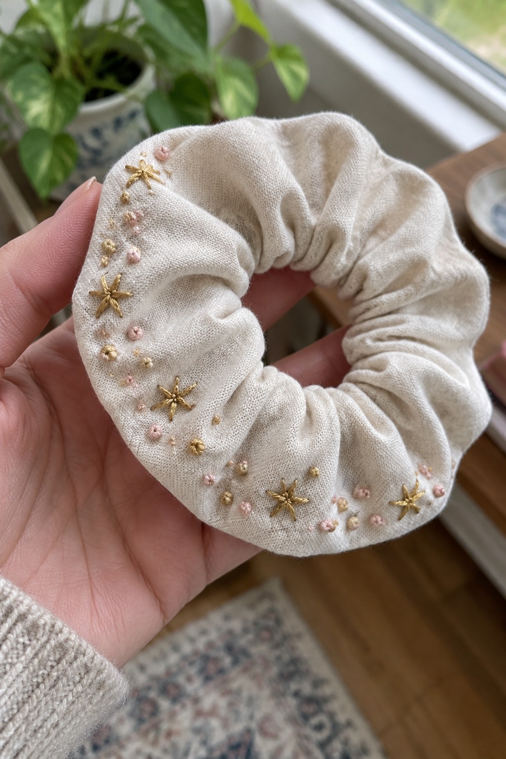 A hand holds a cream gathered scrunchie embroidered with small gold and pink flowers.