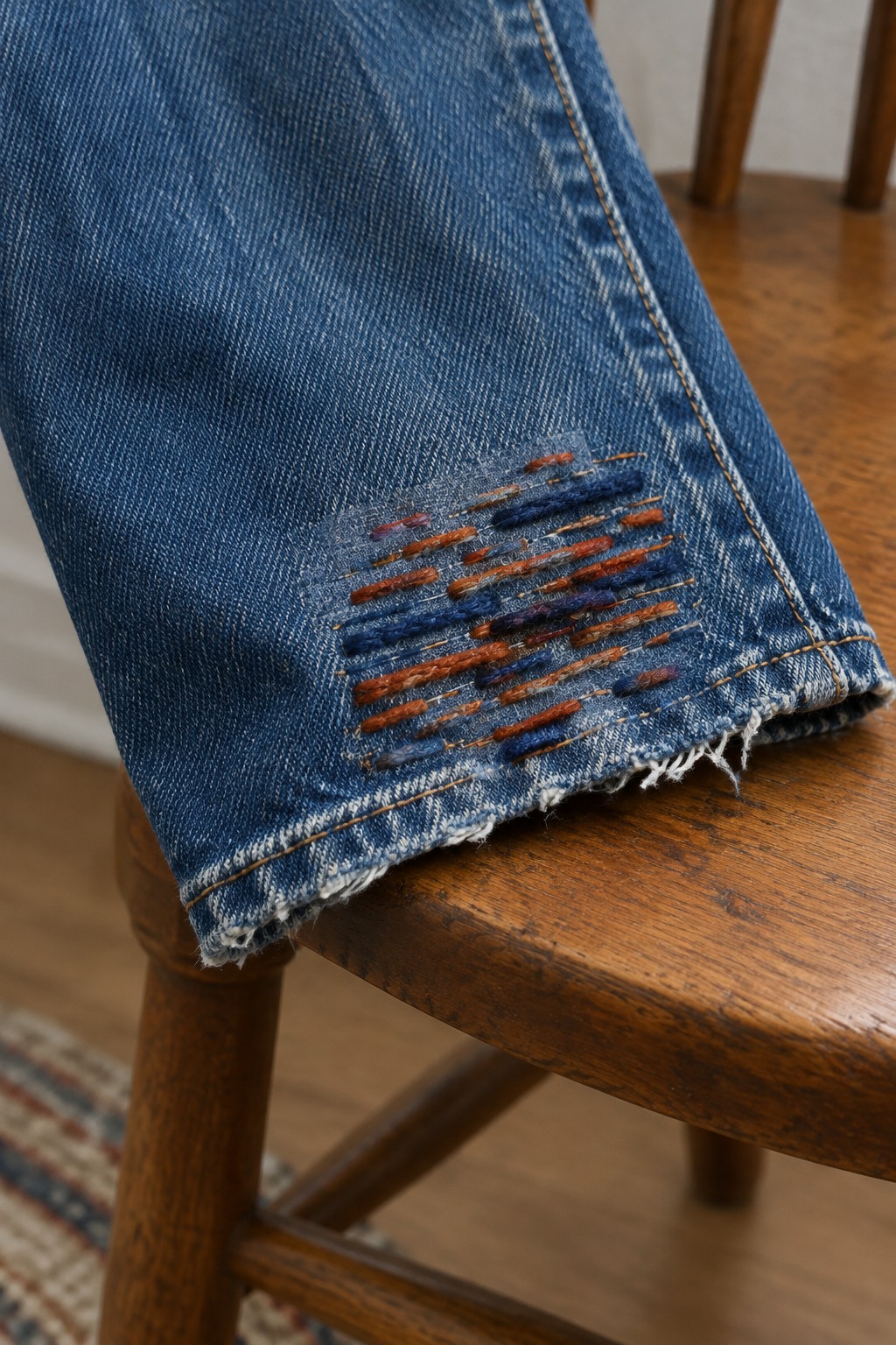 Blue jeans with colorful horizontal stitches above a frayed hem, resting on a wooden chair.
