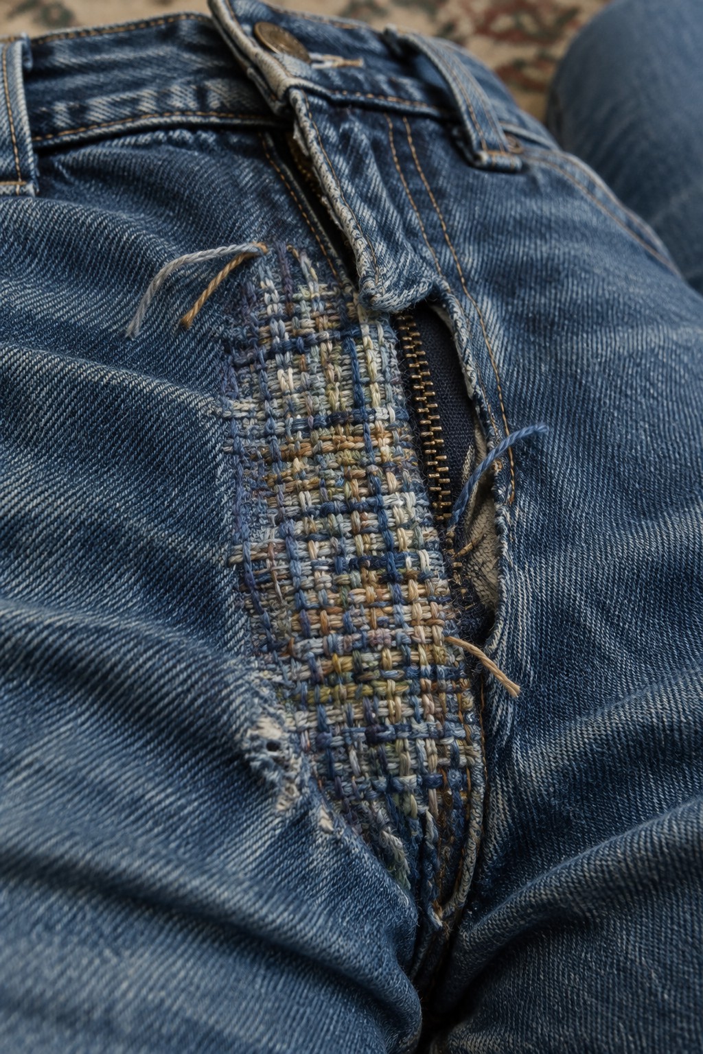 Close-up of blue jeans with a multicolored woven-thread repair beside the zipper.
