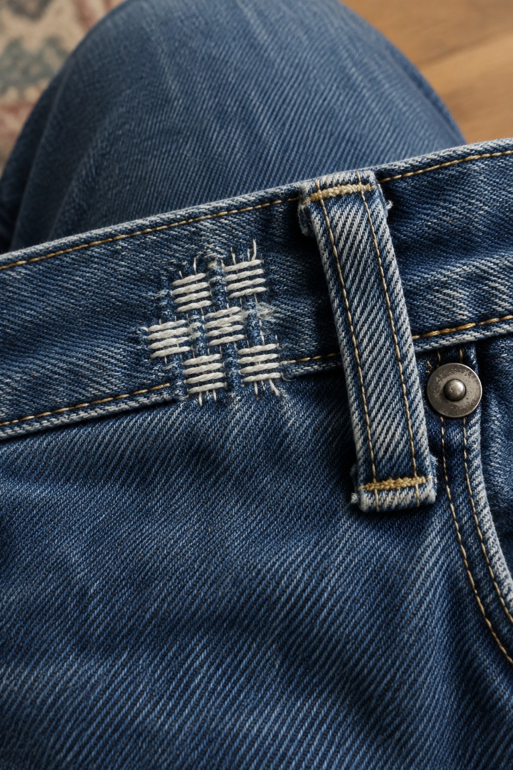 White woven embroidery reinforces a worn area of a blue jean waistband beside a belt loop.