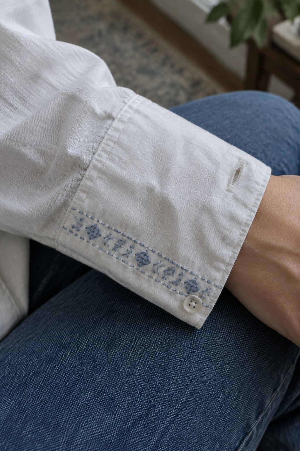 White shirt cuff embroidered with a narrow band of blue geometric stitching and a white button.