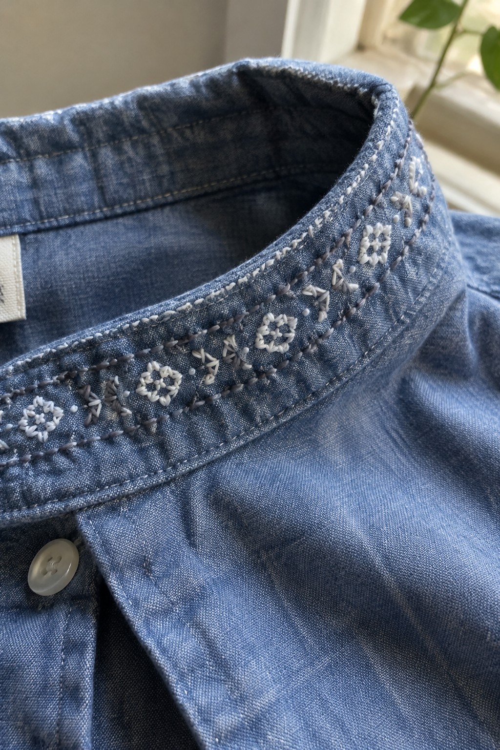 Close-up of a blue denim shirt collar embroidered with white and gray floral motifs and rows of stitches.