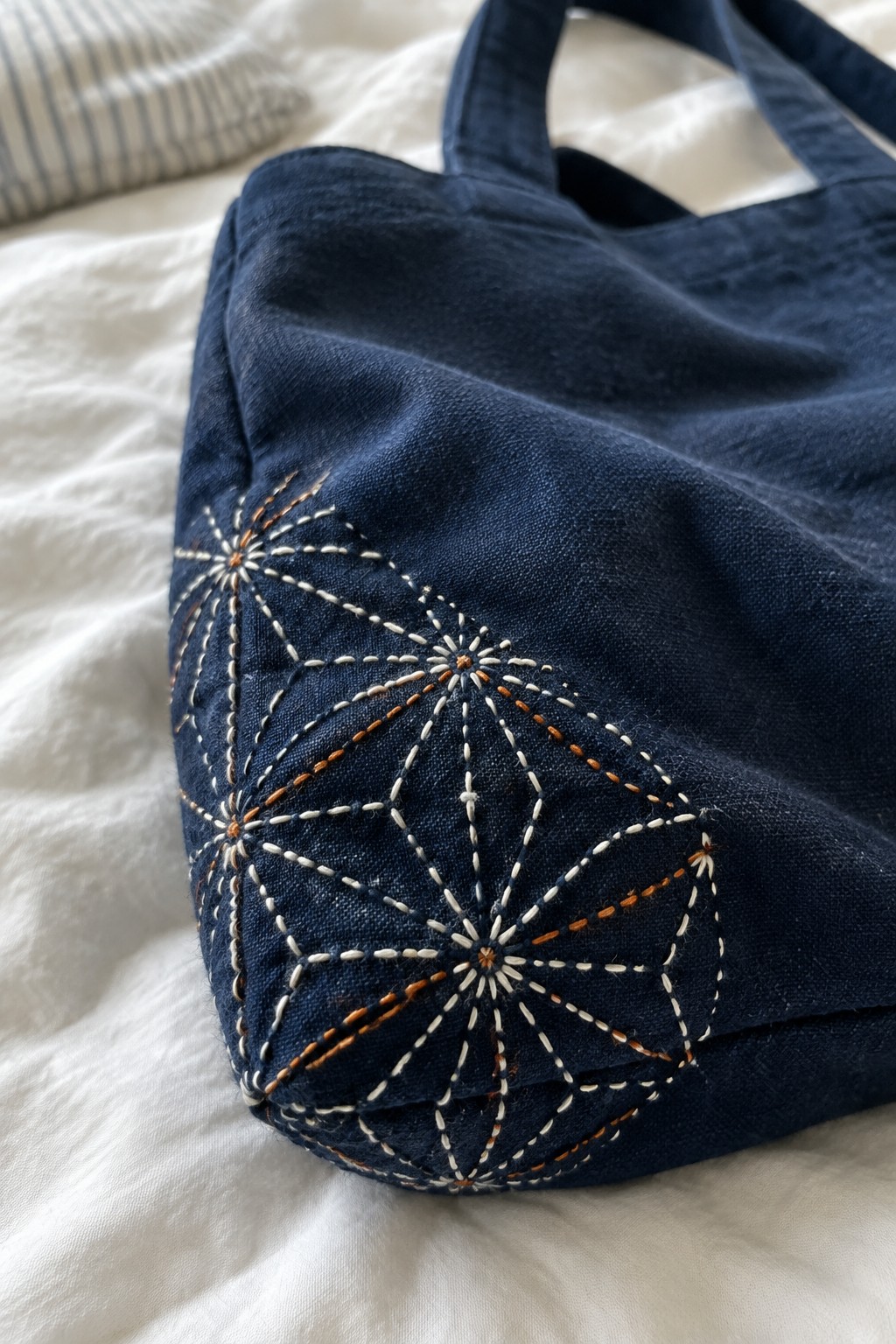 Cream and rust geometric sashiko stitching decorates the corner of a dark indigo tote bag.