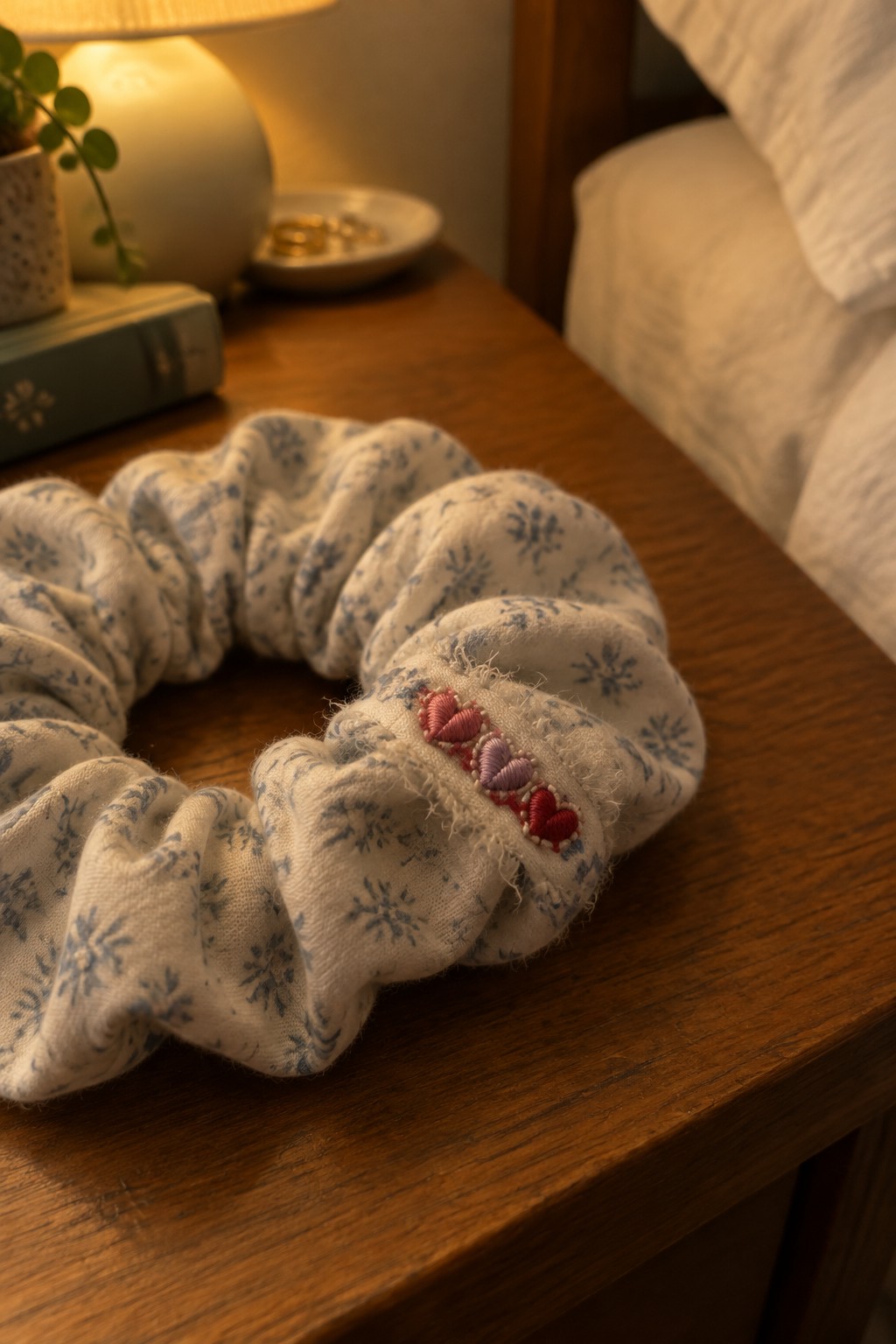 A cream floral scrunchie with small red and pink embroidered hearts rests on a wooden bedside table.