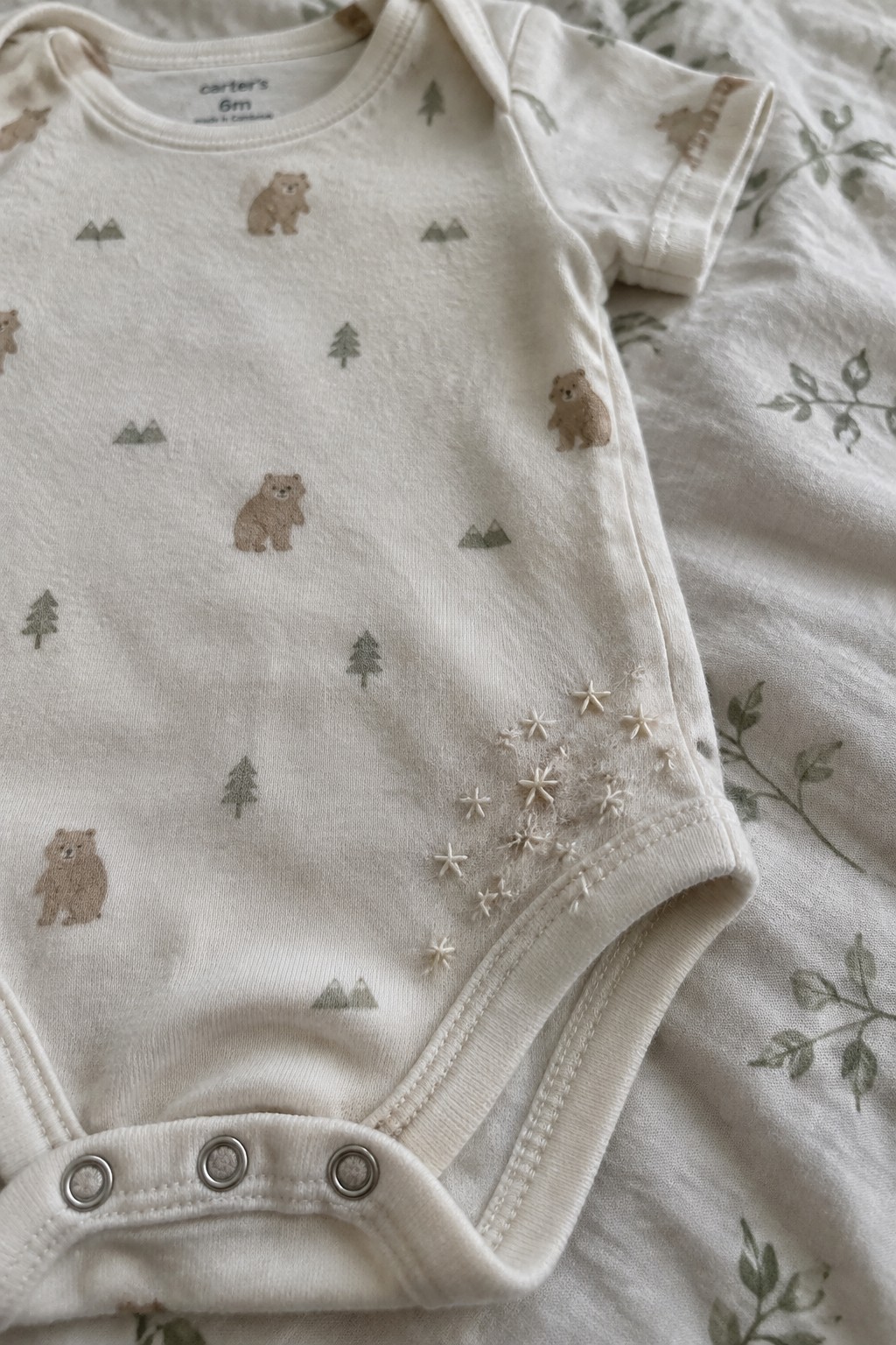 An ivory baby onesie printed with bears, trees, and mountains has a cluster of small white embroidered stars near the leg opening.