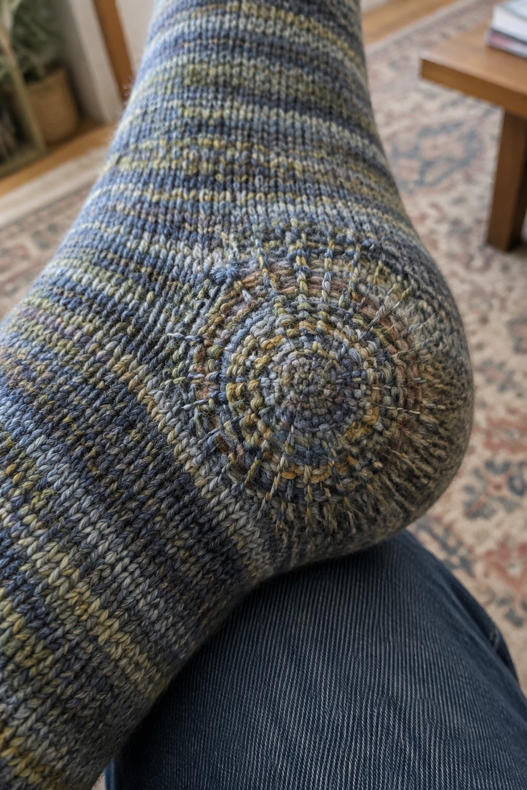 A knitted sock heel has a circular repair stitched in muted blue, green, and tan thread.