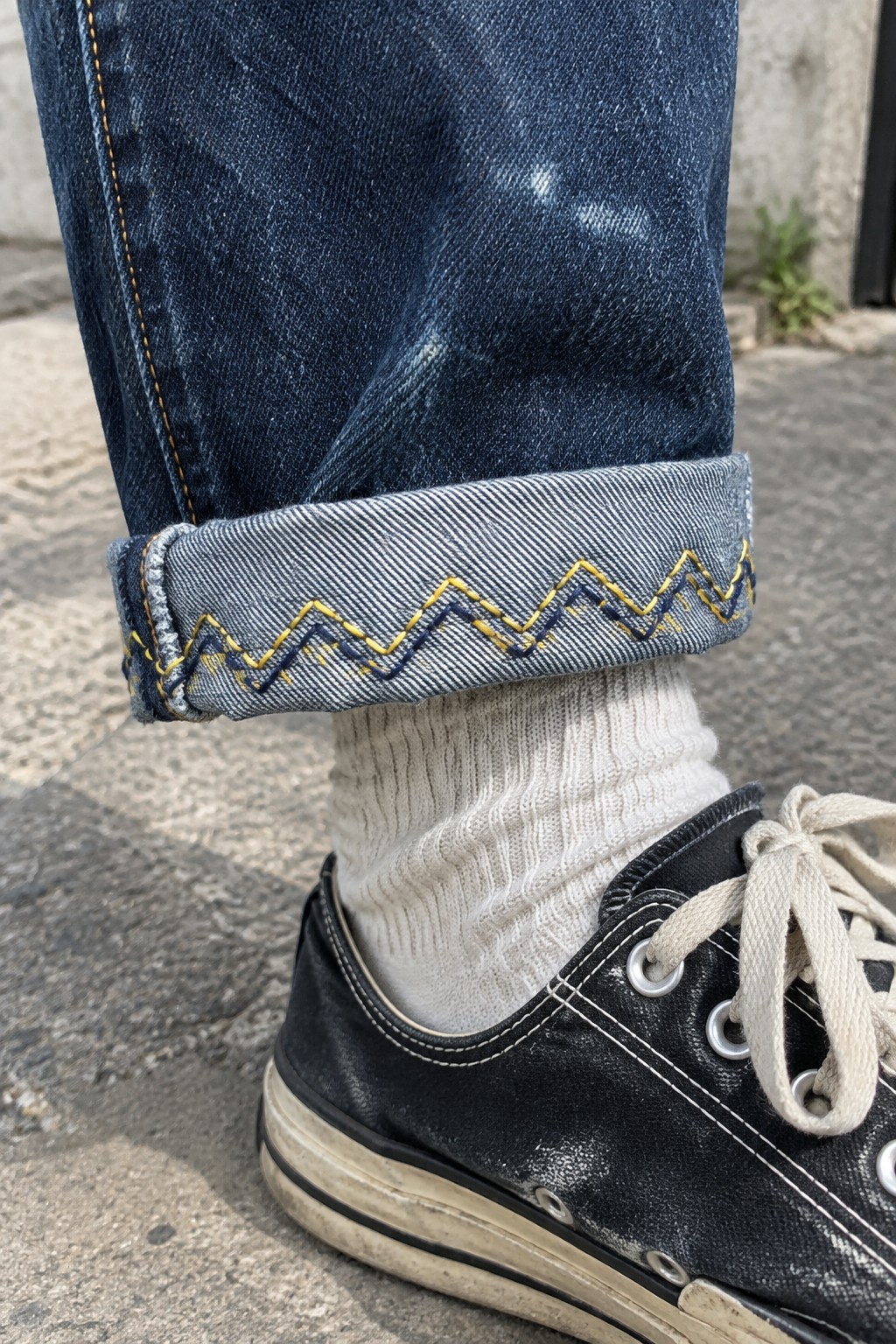 A denim jean cuff with yellow zigzag stitching above a white sock and black canvas sneaker.