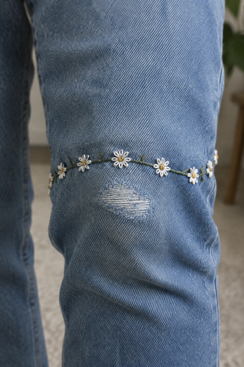 Blue jeans with a small white daisy vine embroidered around the knee above a distressed patch.