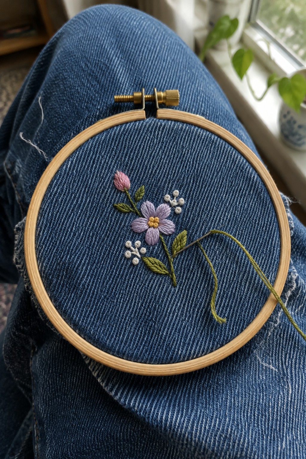 A wooden embroidery hoop holds blue denim with a small embroidered bouquet of purple and pink flowers, white buds, and green stems.